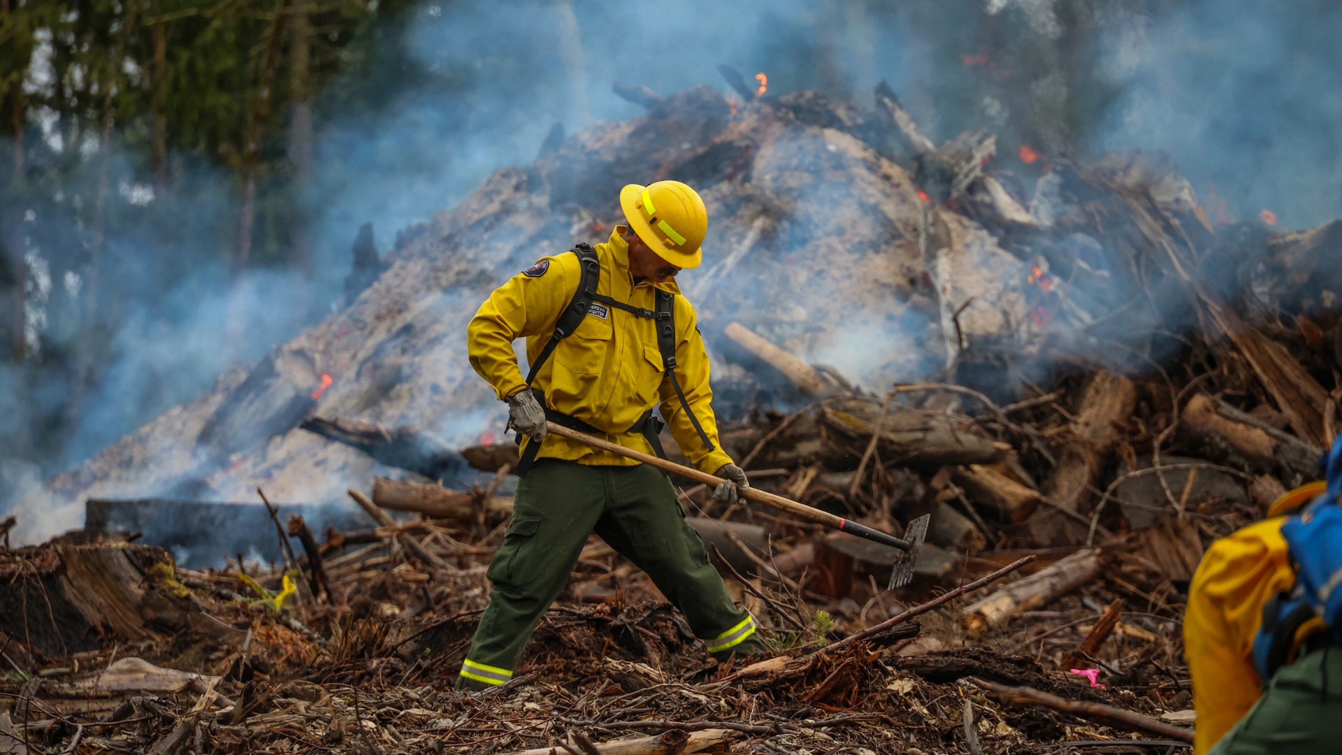 Firefighter in yellow gear uses a tool to manage a smoky, smoldering pile of debris and burned wood in a forested area.