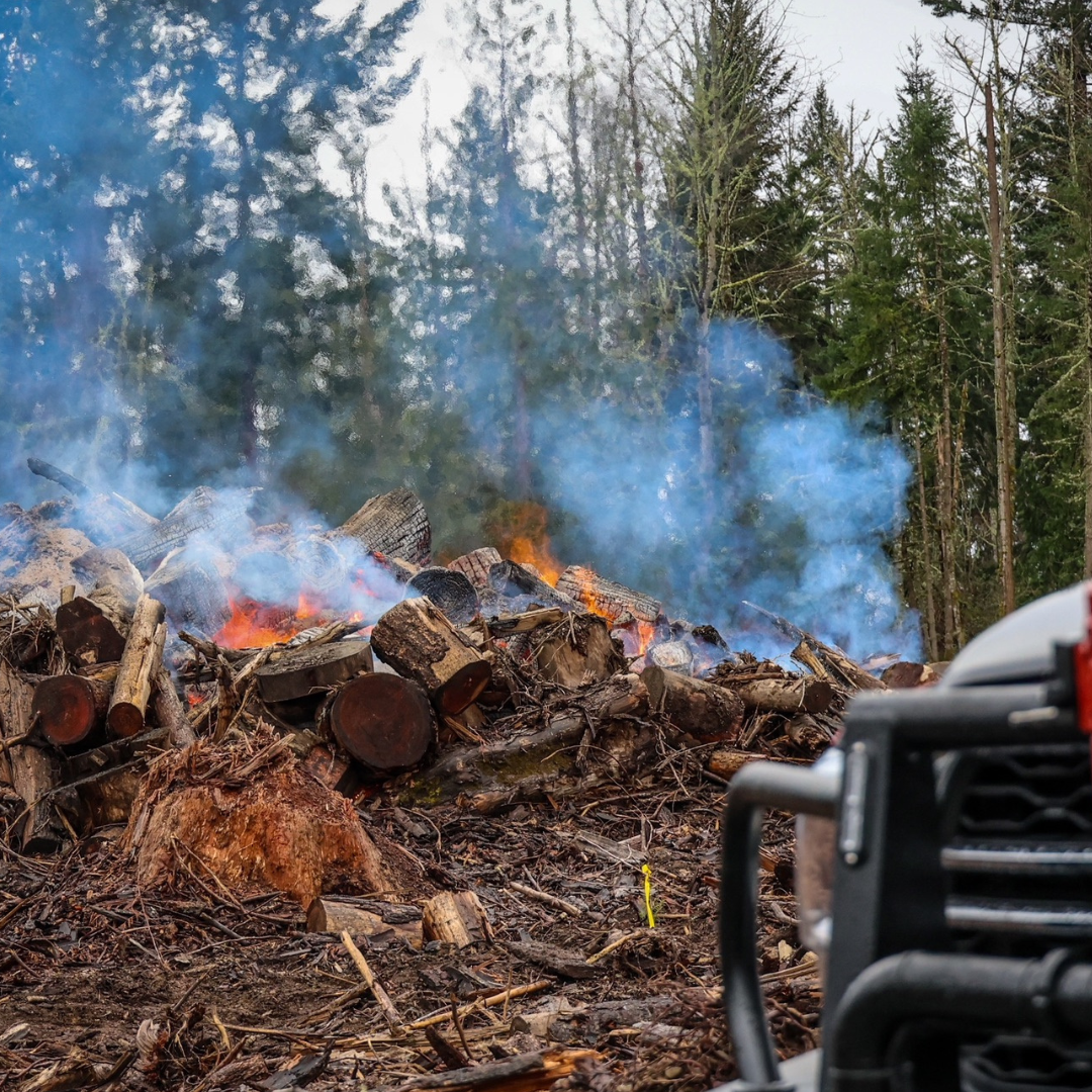 A large pile of burning wood and debris in a forest, with a red fire nozzle mounted on the front of a truck in the foreground.