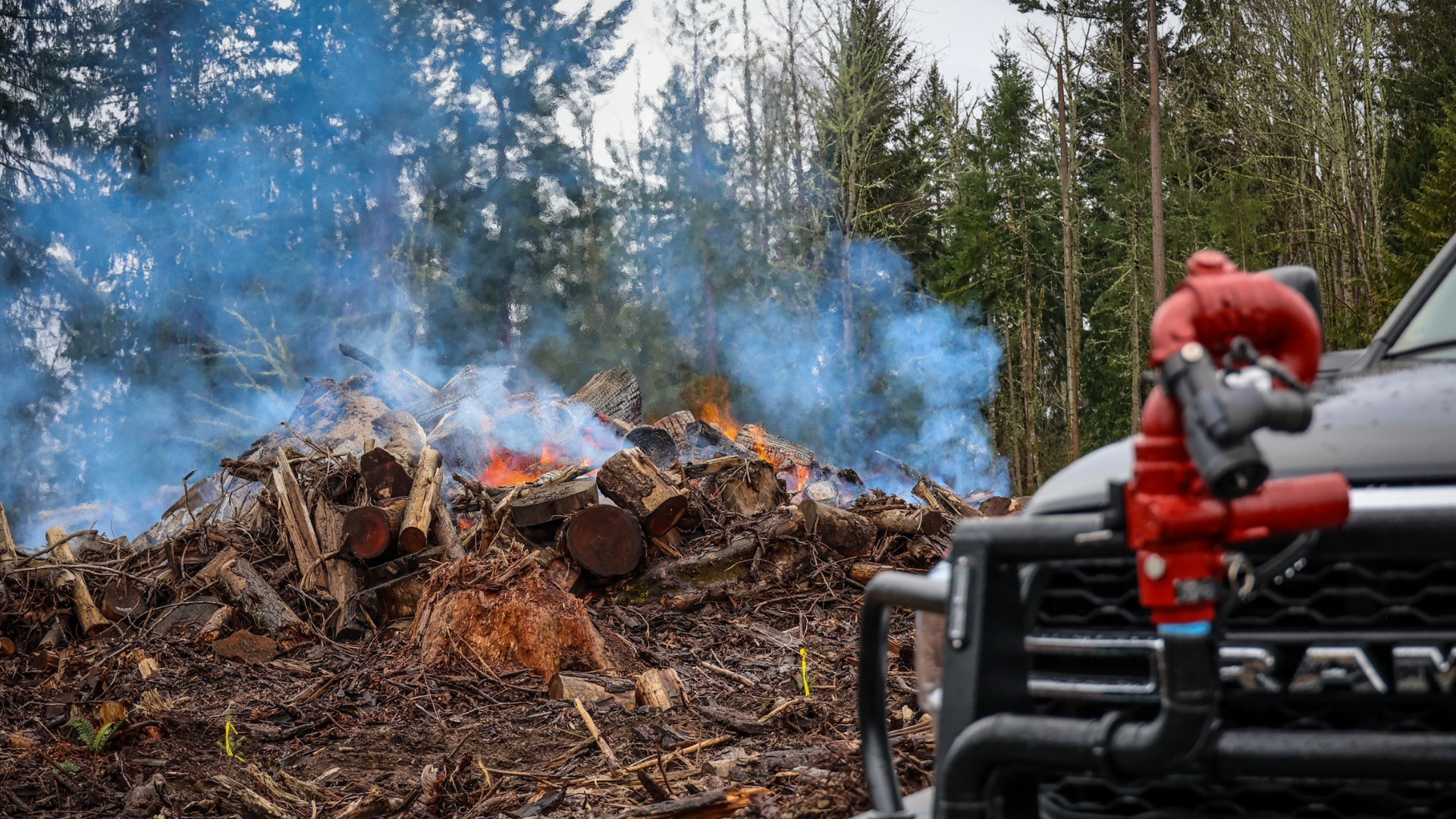 A large pile of burning wood and debris in a forest, with a red fire nozzle mounted on the front of a truck in the foreground.