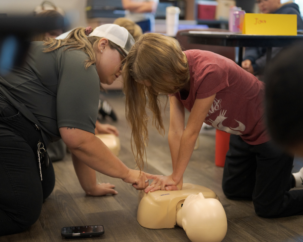 An adult instructs a child on performing CPR chest compressions on a practice dummy while other children observe in the background during a Central Pierce Fire & Rescue Safe Sitter class.