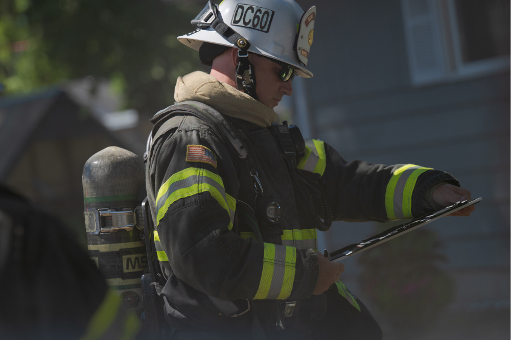 A firefighter in full gear and helmet, reviews a clipboard outside a residential building during daylight.