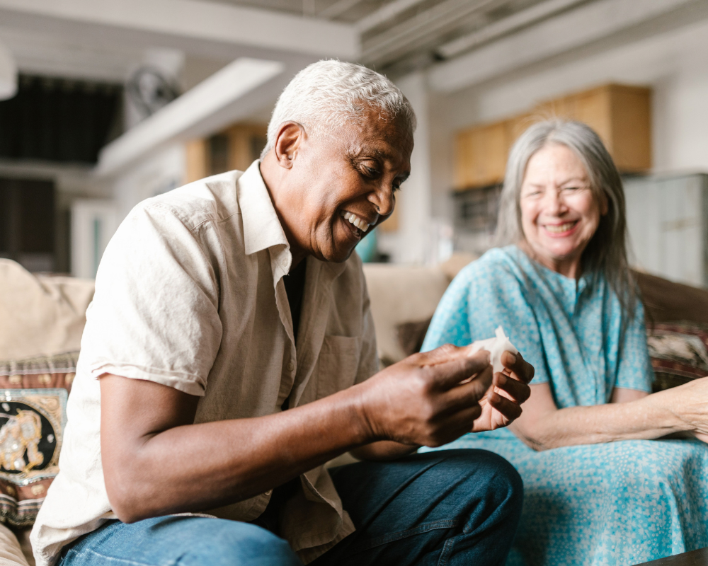 An older man and woman sit on a couch indoors, smiling and laughing together as they discuss Central Pierce Fire & Rescue safety for older adults. A kitchen area is visible in the background.