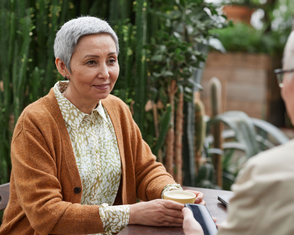 Older woman with short gray hair, in a brown cardigan, enjoys coffee and discusses fall prevention for older adults in an indoor garden setting.